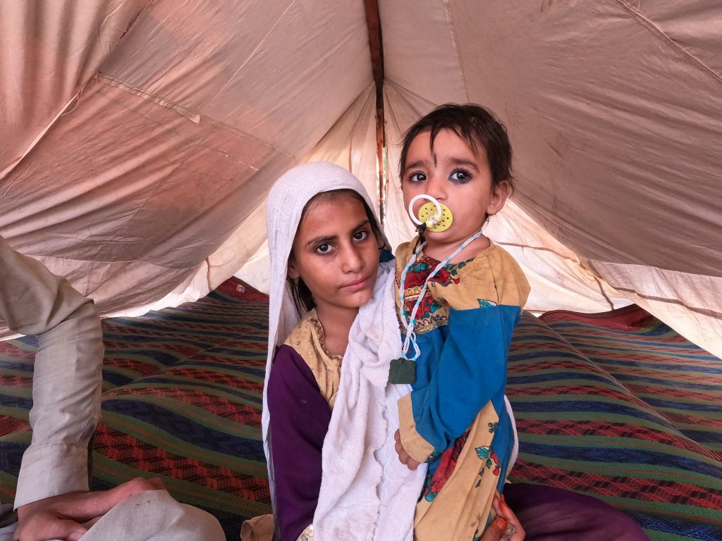Pakistani girl holds her younger sister as they gaze at the camera, in their make-shift tent.